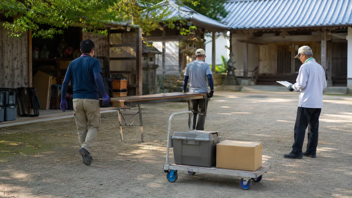 架空の小さな神社の境内で、高齢の氏子を含む3人の当番が台車を使い、机と無地の収納箱を小分けにして運搬表を確認する秋祭り準備の様子